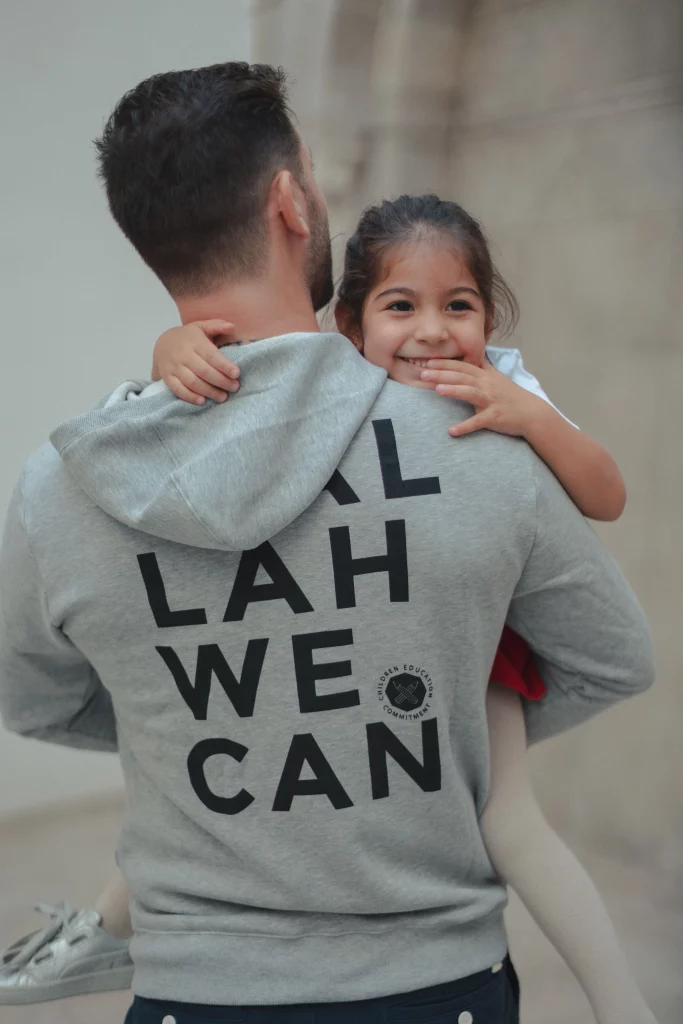 père portant un t-shirt "wallah we can" et tenant son enfant dans ses bras